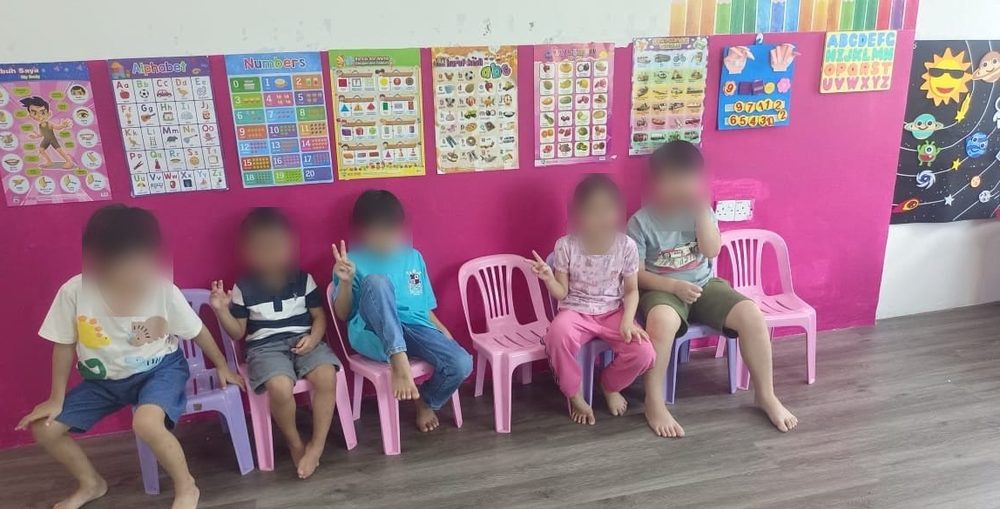 Five children sitting together on small chairs in front of the classroom alphabet and number charts